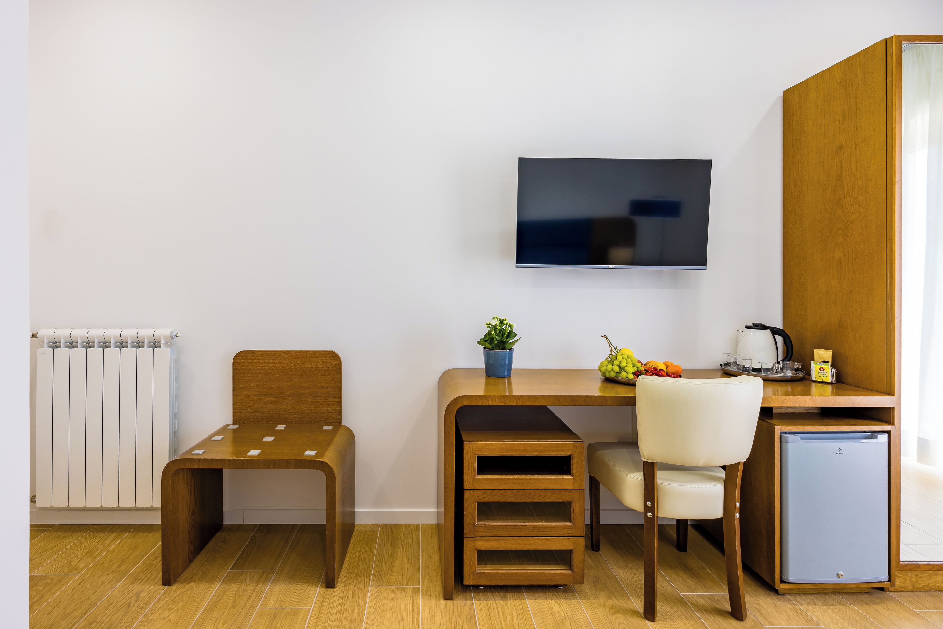 A compact room with a bed, a desk, a chair, a television, a small refrigerator, and a radiator.