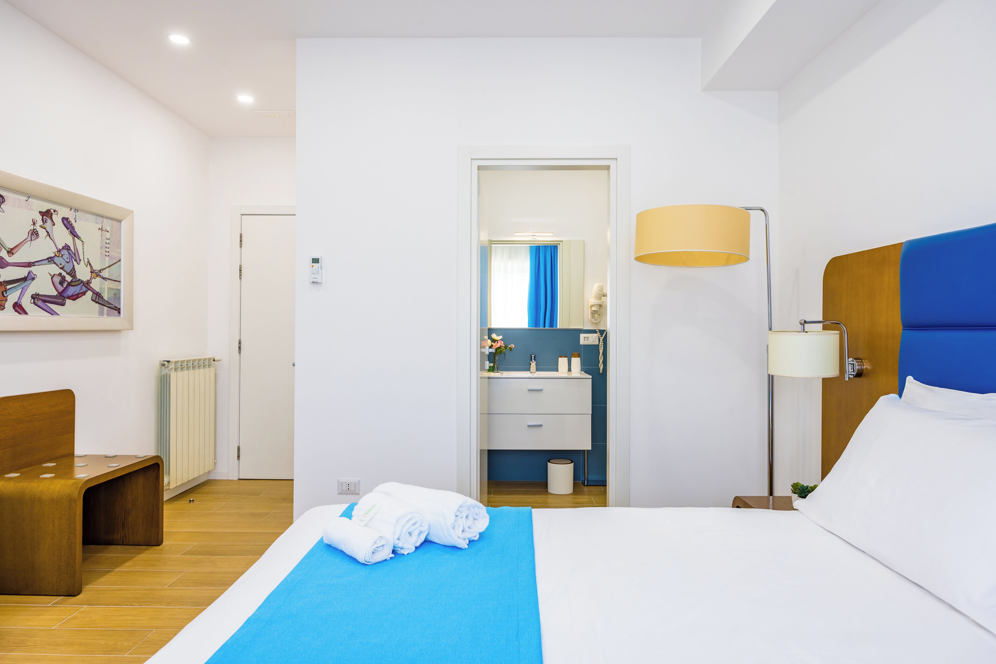A hotel room with a bed, a desk, a chair, a lamp, a bathroom visible through an open door, and a painting on the wall.