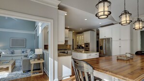 A spacious living room with a large wooden dining table, a kitchen with white cabinetry, and a modern chandelier.