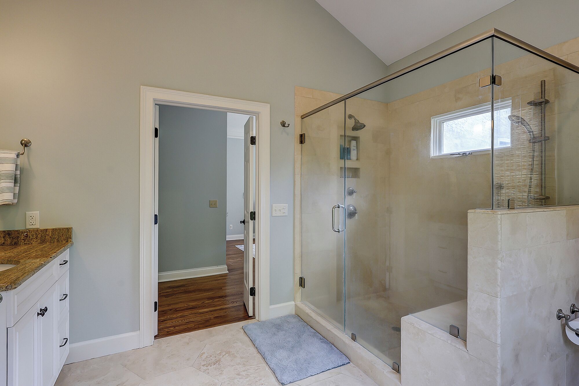 A modern bathroom with a glass shower enclosure, a wooden vanity, and a visible hallway leading to another room.