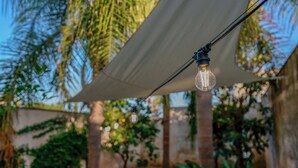 Une guirlande de petites lampes suspendue sous un auvent blanc, dans un décor extérieur agrémenté de verdure et d’un mur de briques en arrière-plan.