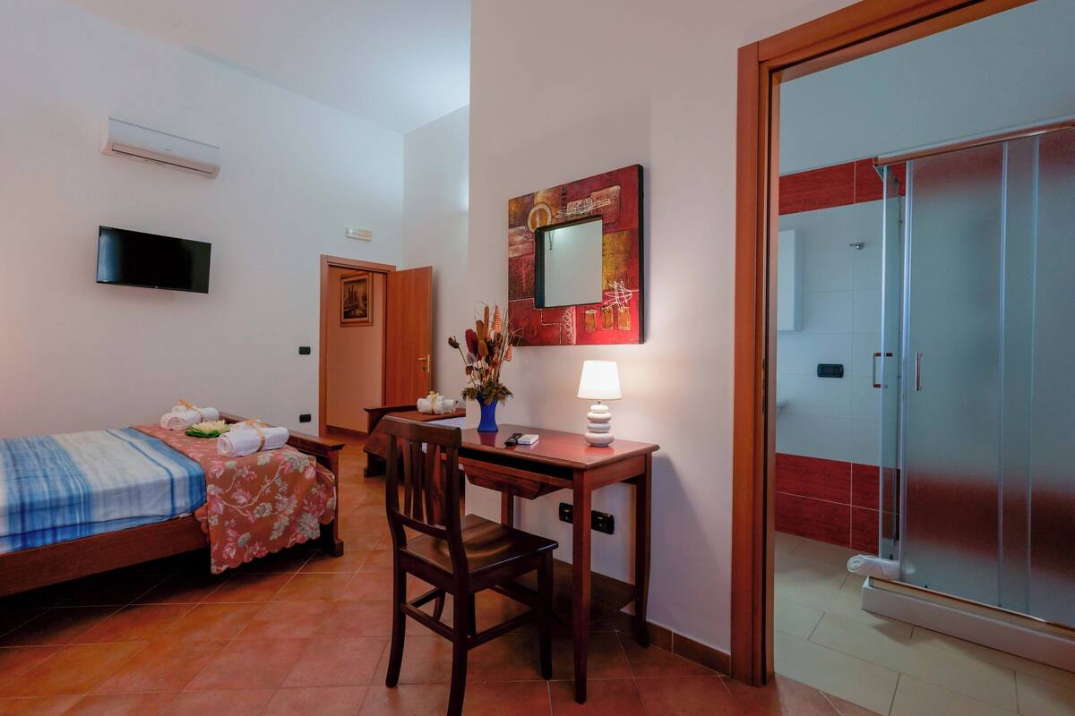 Habitación de hotel con cama, escritorio, silla, televisor, espejo y puerta corredera hacia la zona de ducha.