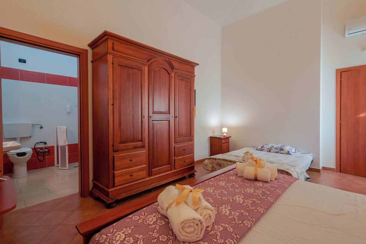 Habitación de hotel con un amplio armario de madera, una cama con ropa de cama estampada, una mesita de noche con lámpara, y un baño visible a través de una puerta abierta.