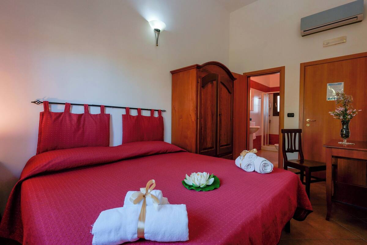 Habitación de hotel con una cama grande, ropa de cama roja, un armario de madera, un escritorio con un jarrón y un baño visible a través de una puerta abierta.