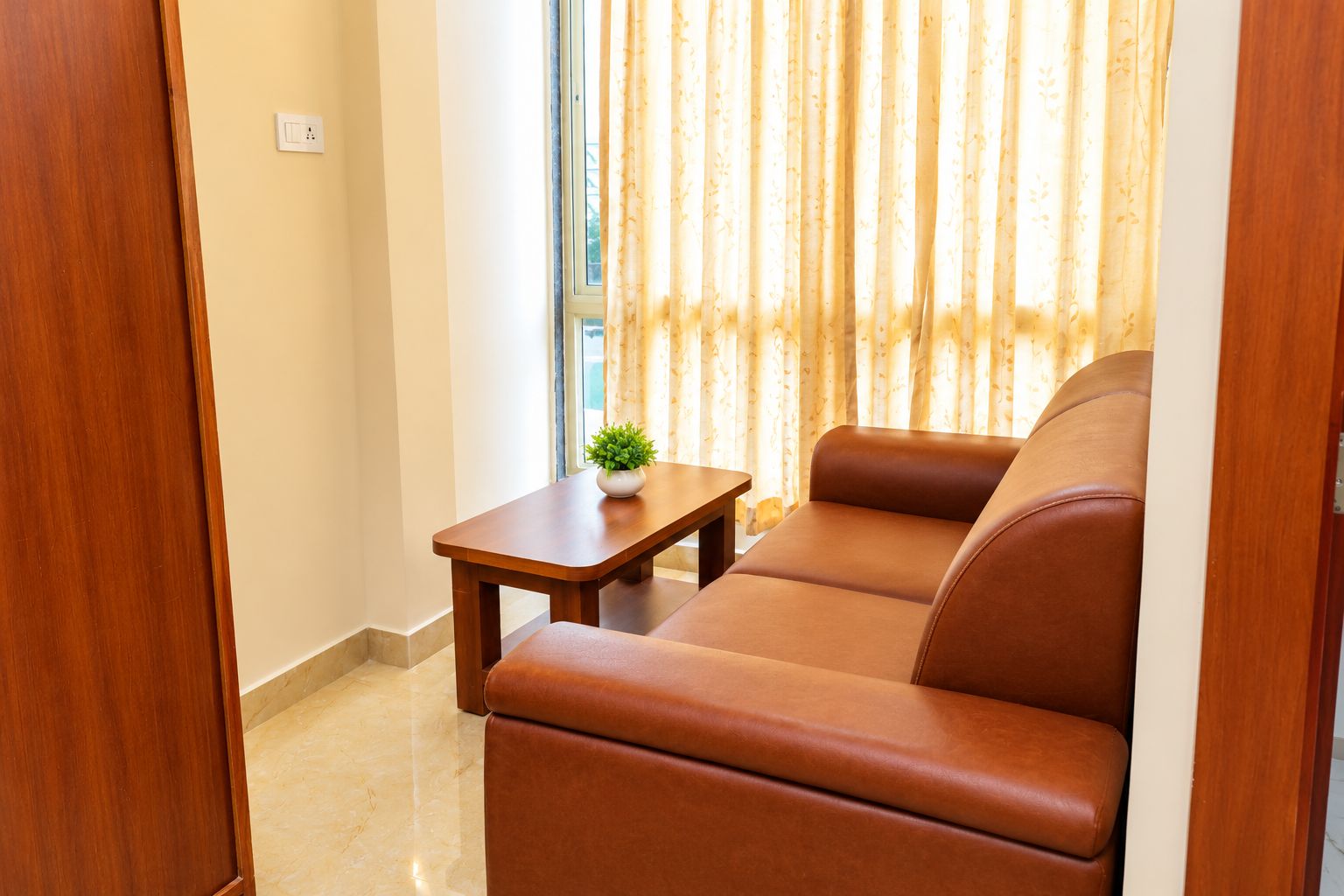 Sala de estar com sofá de couro marrom, uma mesinha de madeira com planta e uma janela com cortinas.