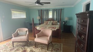 Un dormitorio con una cama, dos sillones, un aparador, un ventilador de techo y una ventana con persianas.