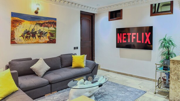 A living room with a grey sofa, a glass coffee table, a painting, a wall-mounted TV displaying 'NETFLIX', and a small plant.
