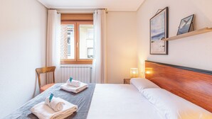 Et hotelværelse med seng, stol, vindue med gardiner, et sengebord med lampe og et billede på væggen.