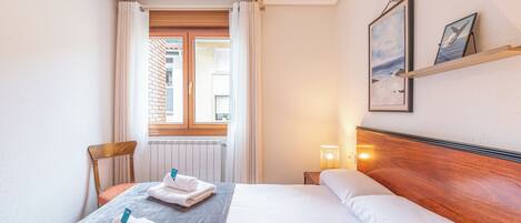 A hotel room with a bed, a chair, a window with curtains, a bedside table with a lamp, and artwork on the wall.
