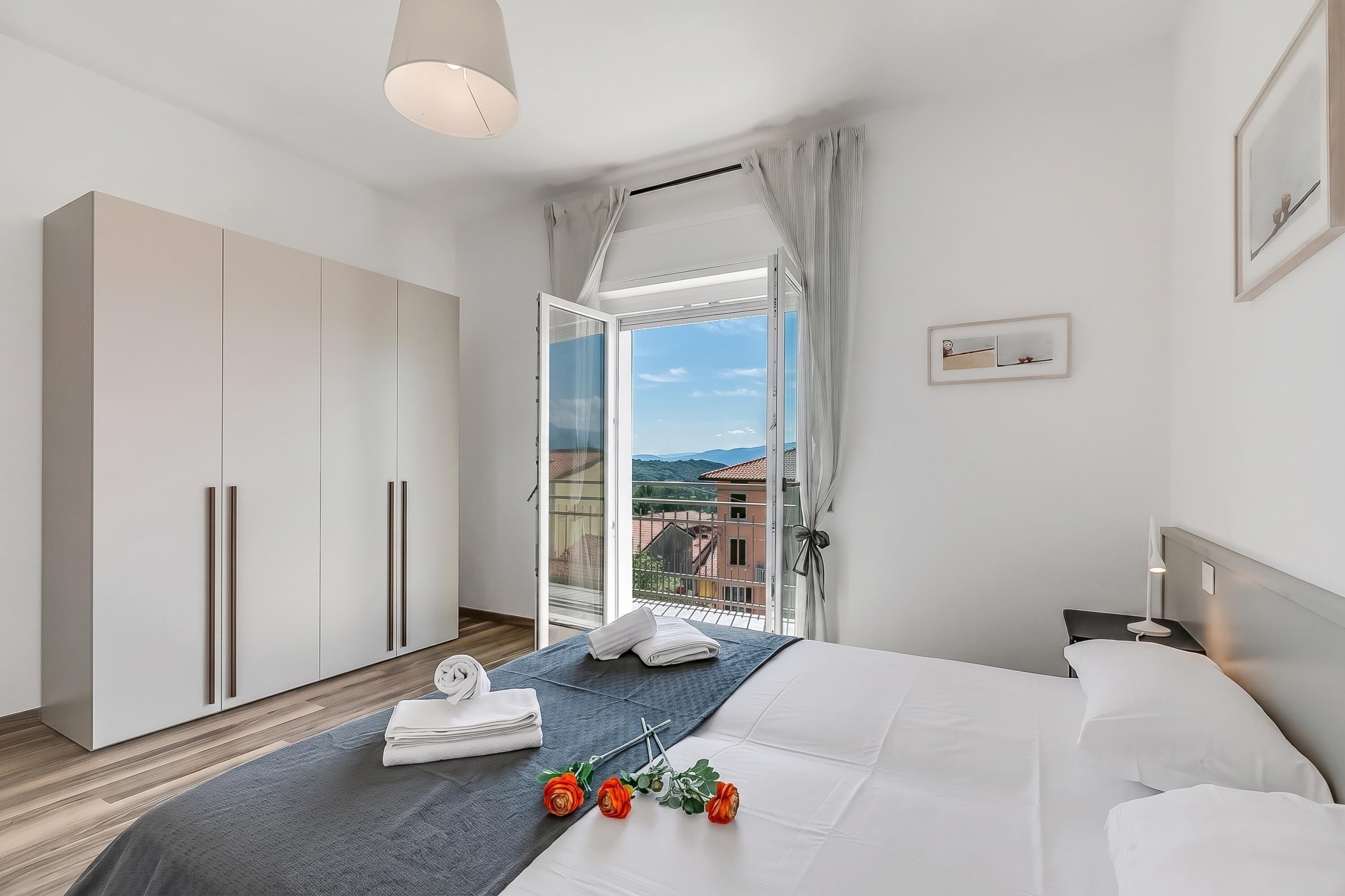 A modern bedroom with a large bed, a wardrobe, a nightstand, and a view of the city through a glass door.