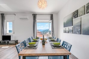 Zona pranzo con tavolo in legno apparecchiato per sei persone, circondato da sedie con schienali traforati, una televisione su un mobile e vista su un paesaggio montuoso attraverso un'ampia finestra.