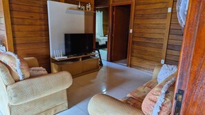 Ein gemütliches Wohnzimmer mit einem Flachbildfernseher auf einem Holzboden, zwei beigefarbenen Sesseln und einem kleinen Tisch mit einer Fernbedienung.