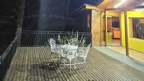 Eine Holzterrasse mit Metallstühlen und einem Tisch, umgeben von Bäumen und einem Gebäude mit gelber Fassade.