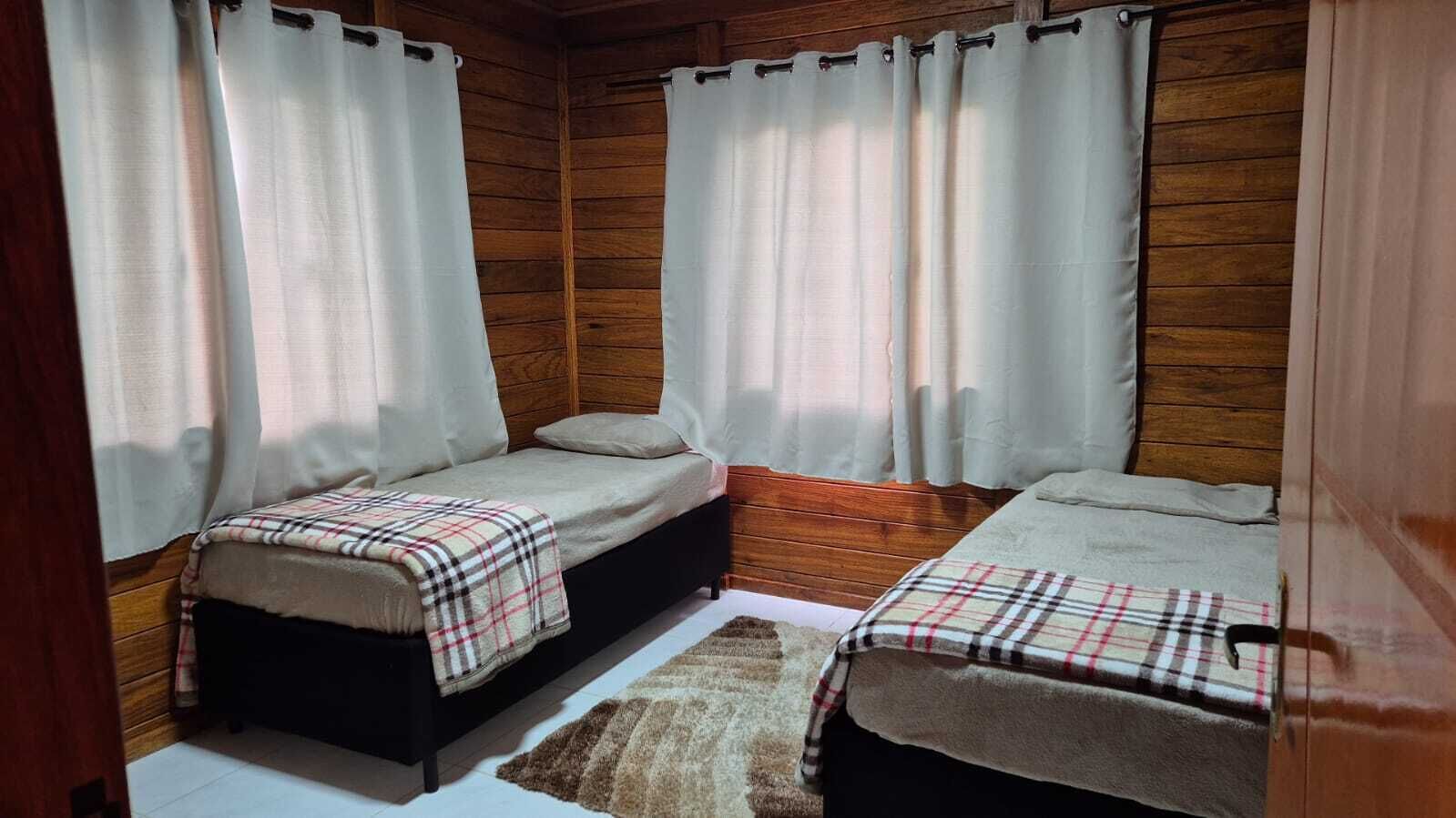 Una habitación con dos camas individuales, paredes de madera, cortinas blancas y una alfombra marrón.