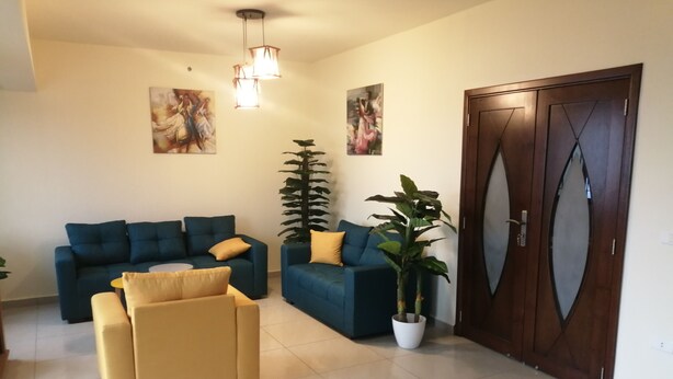A living room with a blue sofa, a yellow armchair, and a large potted plant. There are two paintings on the wall and a wooden door with a glass panel. - Lovely Appartment in Daroun. Kesserwan (kesrwan)