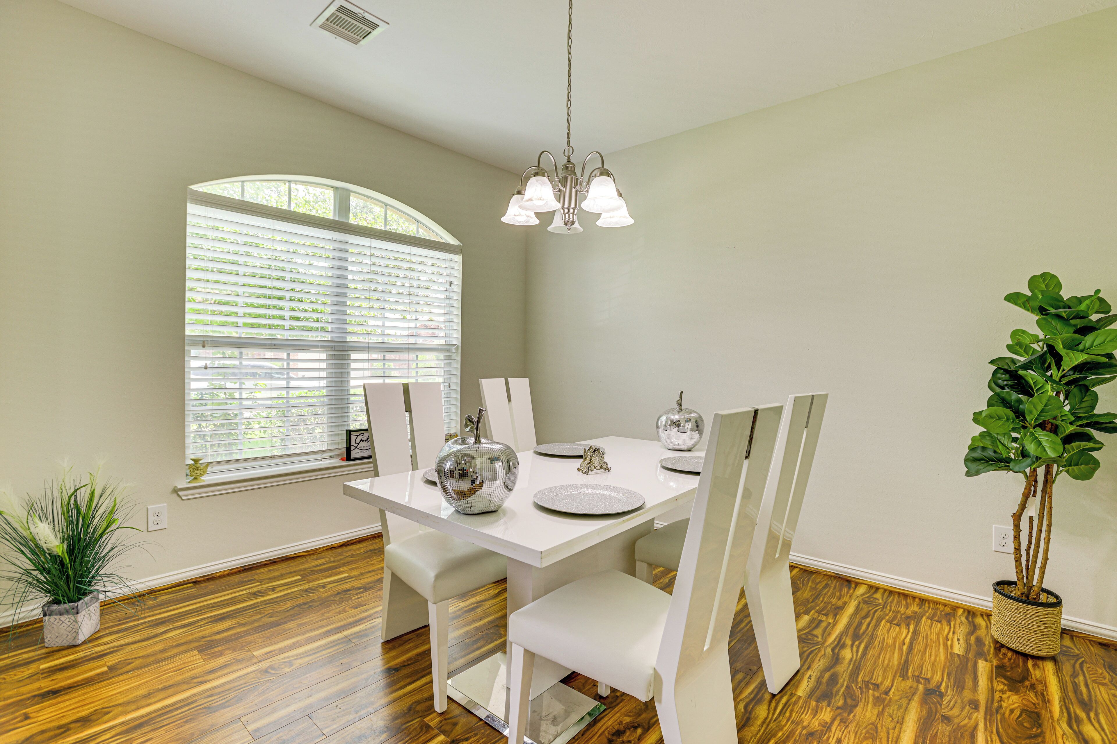 A dining area with a white table, chairs, and a chandelier. There is a window with blinds, a potted plant, and a wooden floor.
