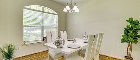 A dining area with a white table, chairs, and a chandelier. There is a window with blinds, a potted plant, and a wooden floor.