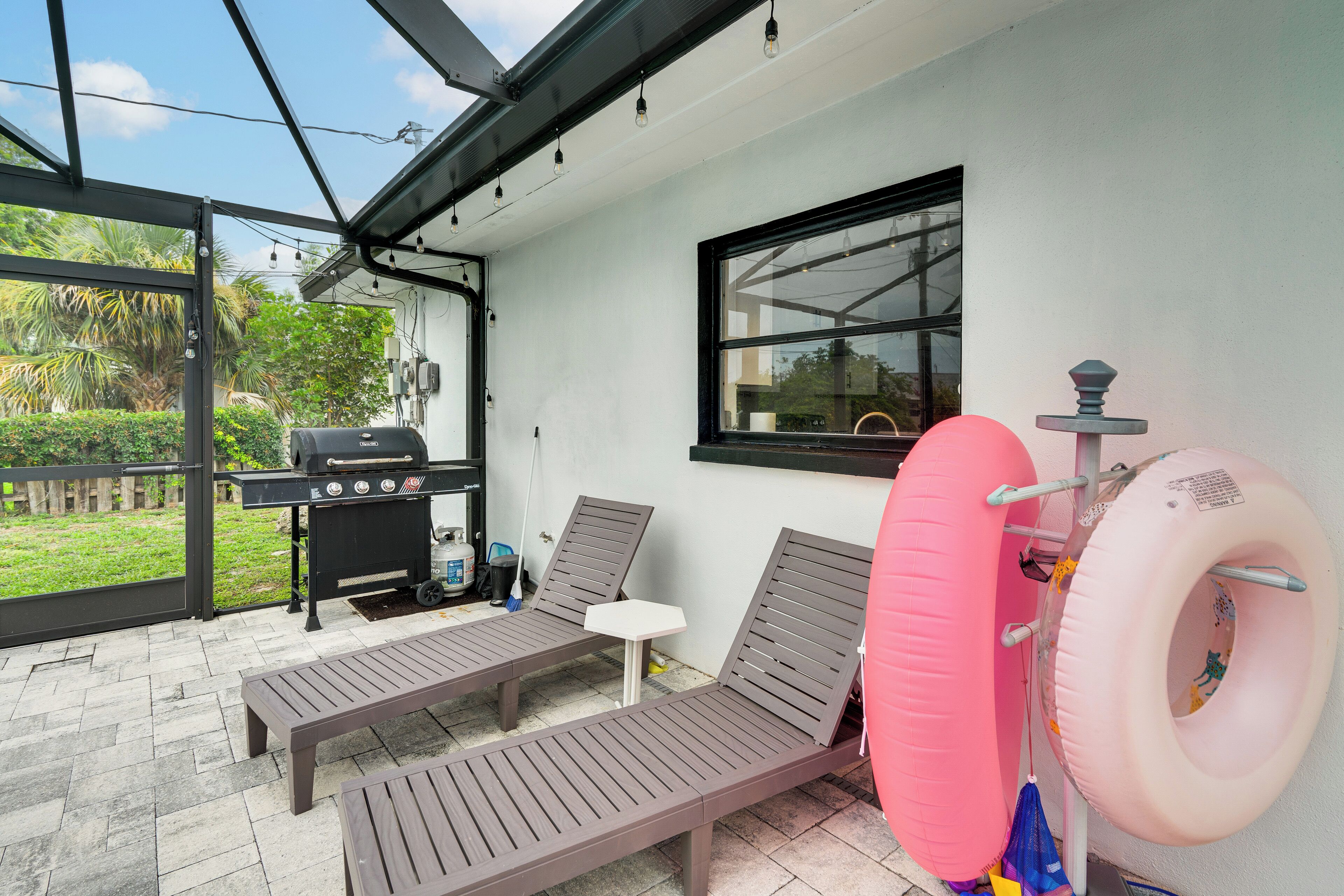 An outdoor patio with two lounge chairs, a small table, a grill, and a window overlooking greenery.