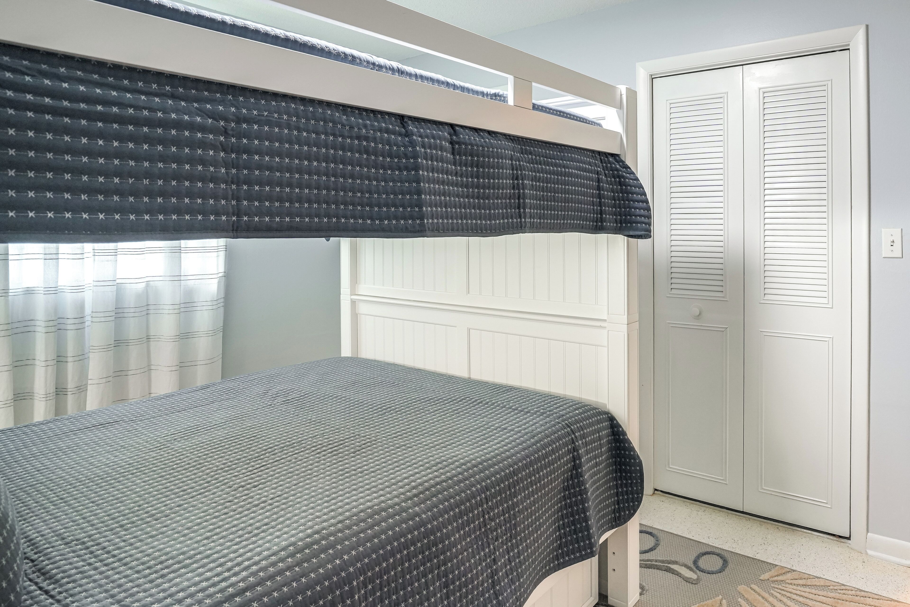 Un llit superposat amb un estovat estampat, una finestra amb cortines i una porta blanca amb manilla.
