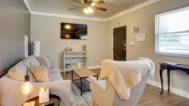 A living room with a sofa, armchair, ceiling fan, and a large window. - 3 Mi to Shops & Eats in Uptown: Greenwood Studio (Greenwood)