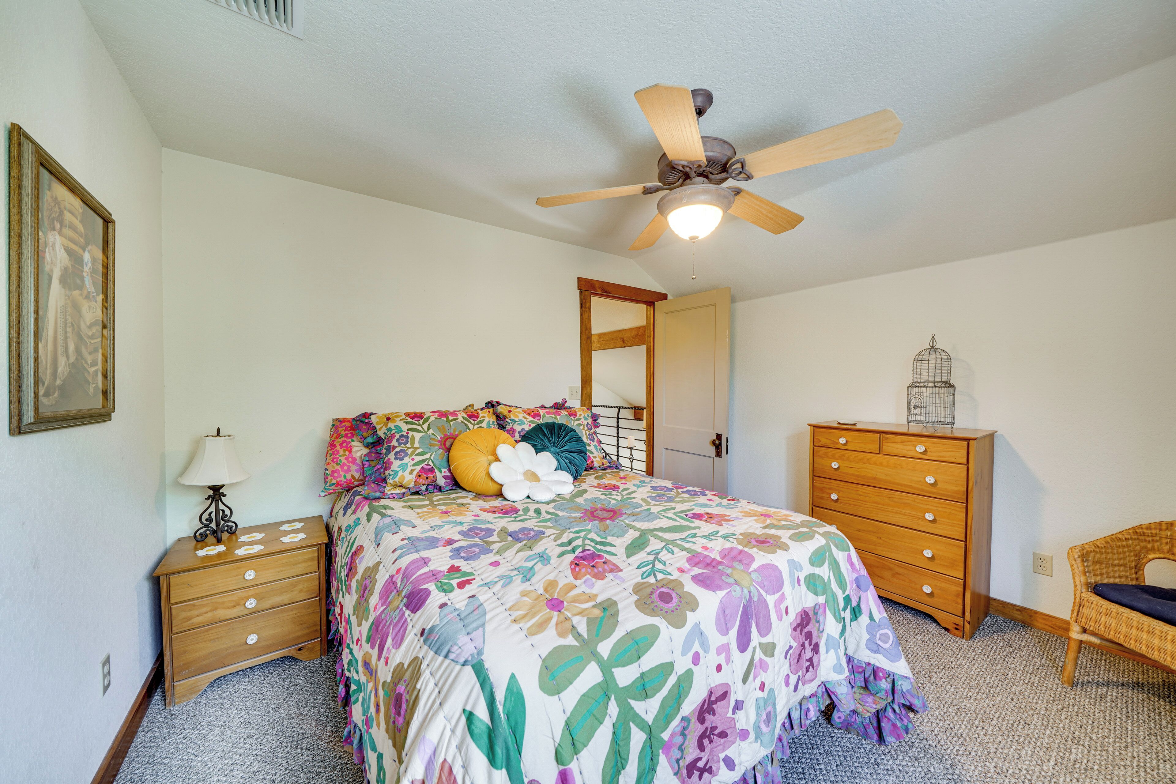 A bedroom with a bed, a wooden dresser, a chair, a ceiling fan, and a door leading to another room.