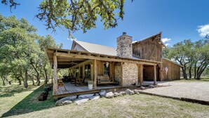 A rustic wooden cabin with a stone chimney, a covered porch with seating, and an American flag on the porch. - Serene Hill Country Home! Hike, Sleep, Unplug! (Kerrville)