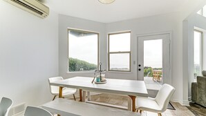 Un área de comedor con una mesa y sillas blancas, ventanas amplias y vistas al exterior.