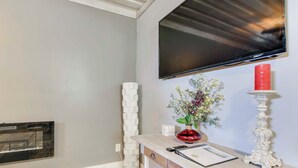 Ein modernes Wohnzimmer mit einem an der Wand befestigten Flachbildfernseher, einem weißen Konsolentisch mit Blumen in einer Vase, einem roten Kerzenhalter und einer dekorativen weißen Kerze.