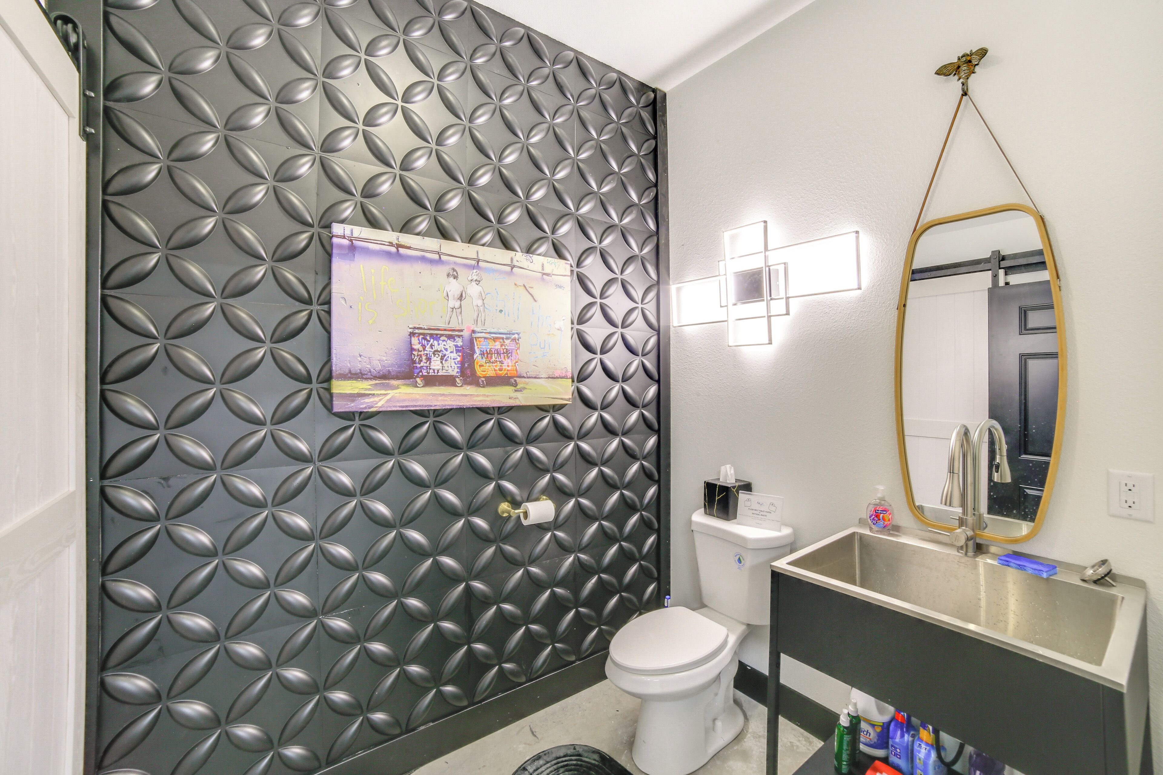 A bathroom with a patterned wall, a toilet, a sink with a mirror, and a trash bin.