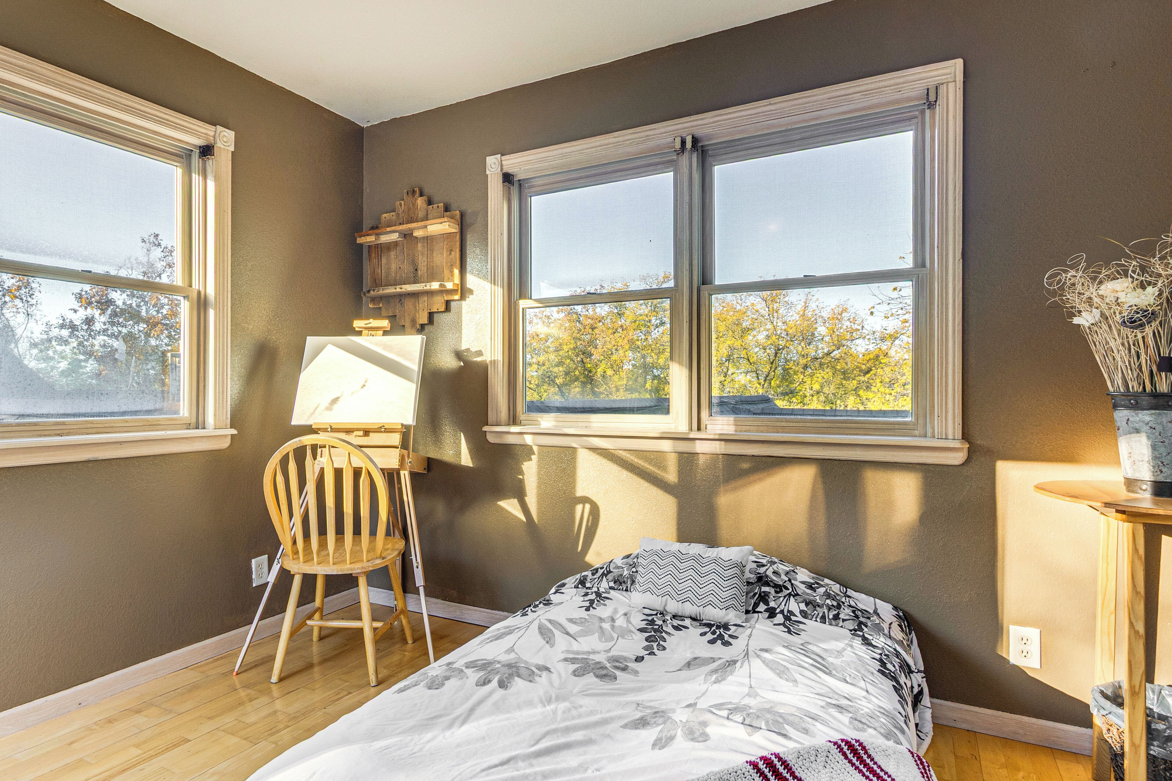A bedroom with a bed, two windows, a desk with a chair, and a small table with a vase.