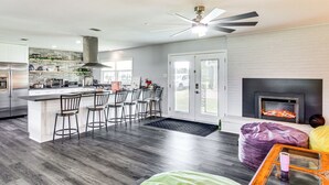 A modern kitchen with a central island, bar stools, a fireplace, and a dining area with a view of the outdoors.