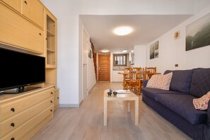 Un salon avec un téléviseur, des meubles en bois, un canapé, une table basse et, au fond, une cuisine.