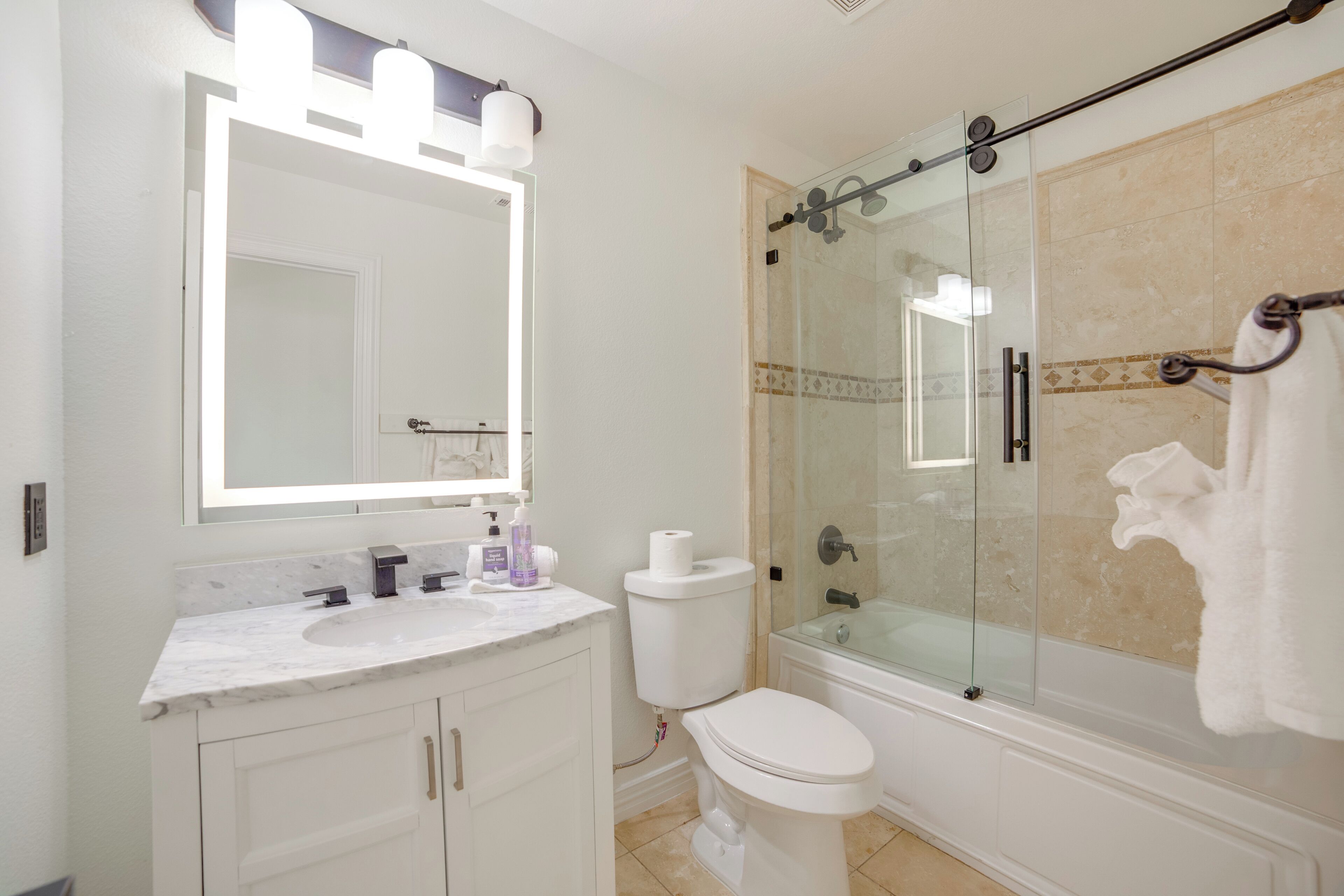A bathroom with a glass-enclosed shower, a white toilet, a marble sink, and a large mirror with lights.