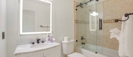 A bathroom with a glass-enclosed shower, a white toilet, a marble sink, and a large mirror with lights.