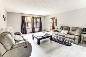 Una sala de estar moderna con un sofá gris, una mesa de centro negra y un sofá beige. Hay dos ventanas con cortinas, una alfombra pequeña y un mueble para televisor.