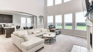 A spacious living room with a sectional sofa, a round coffee table, and a fireplace. There is a kitchen area with dark cabinets and a dining table with chairs. Large windows provide natural light and views of the outdoors. - Grand Family Home w/ Patio & Fenced Yard in Arcola (Rosharon)