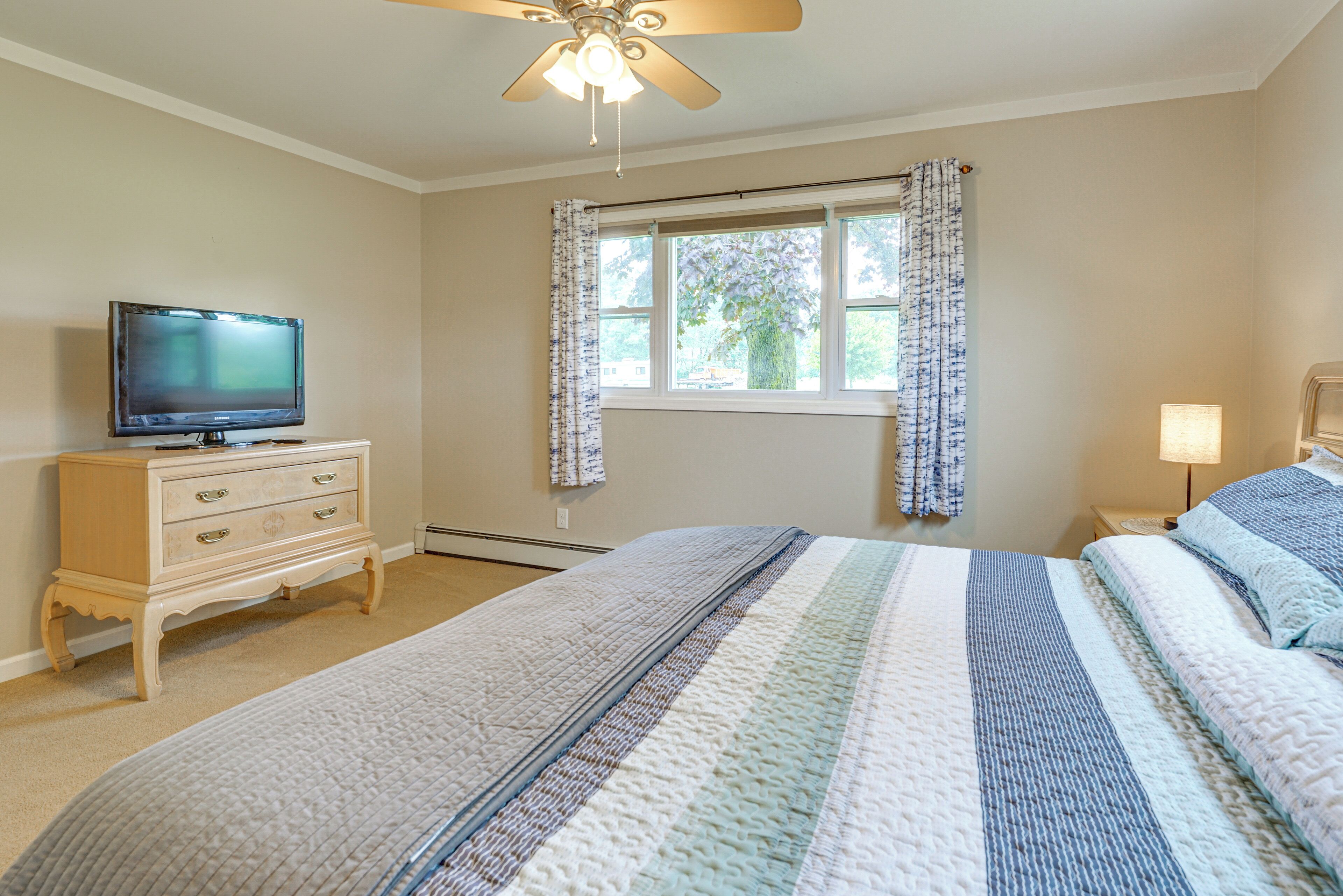 Een slaapkamer met een bed, een dressoir, een televisie, een raam met gordijnen en een plafondventilator.