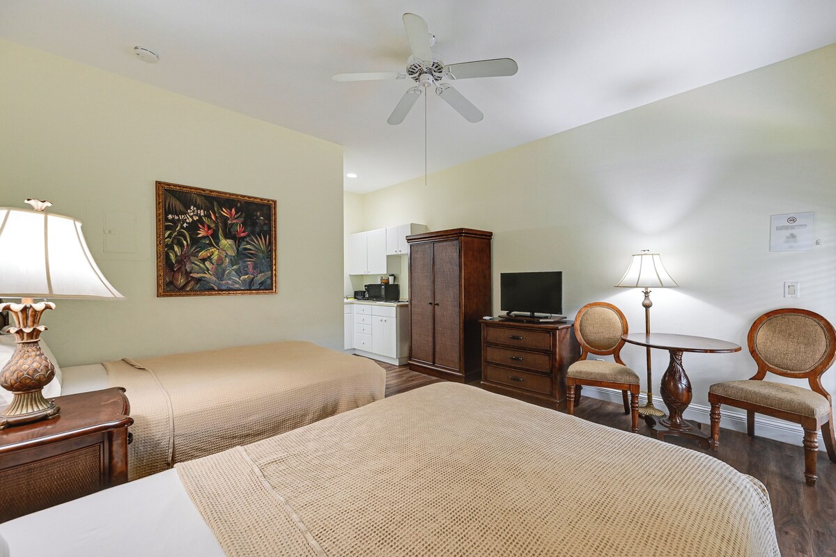 A hotel room with two beds, a ceiling fan, a painting, a desk with a lamp, a television, and a dining area with chairs and a table.