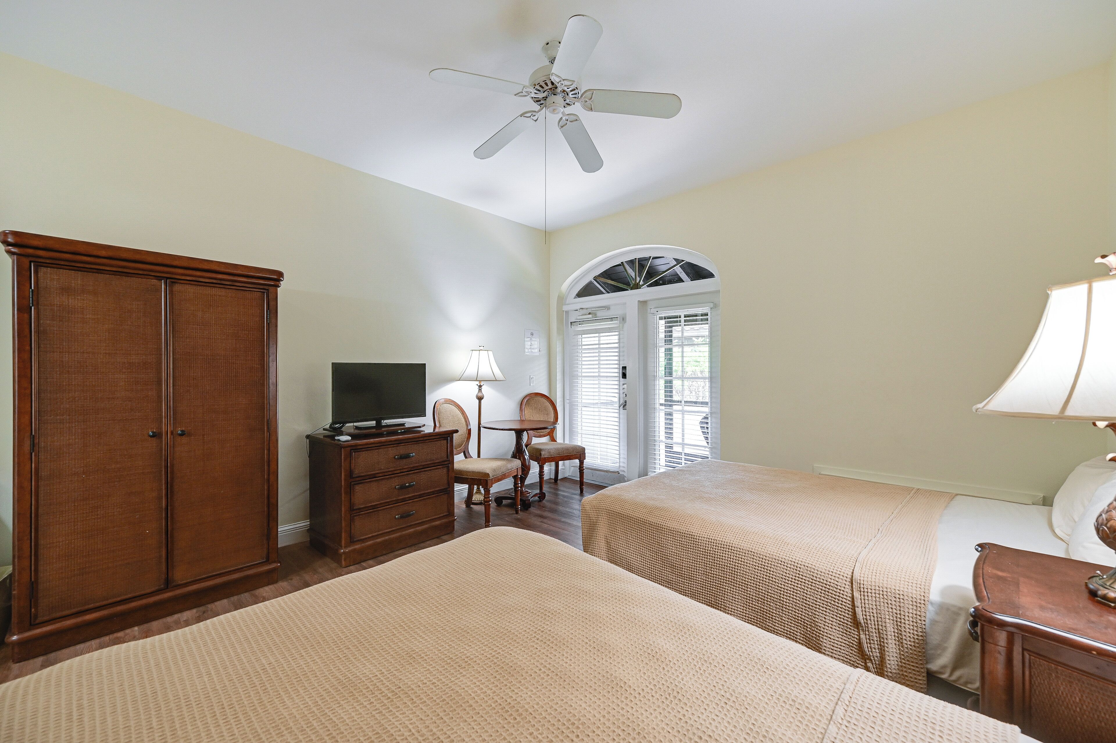 A hotel room with two beds, a wooden wardrobe, a desk with a lamp, a television, and a ceiling fan.