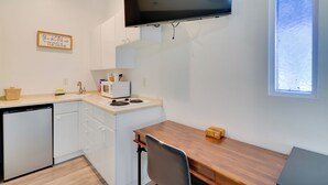 Cocina compacta con estufa integrada, microondas y lavavajillas. Cuenta con una mesa de comedor de madera con una silla, un ventanal pequeño y un televisor de pantalla plana montado sobre la encimera.