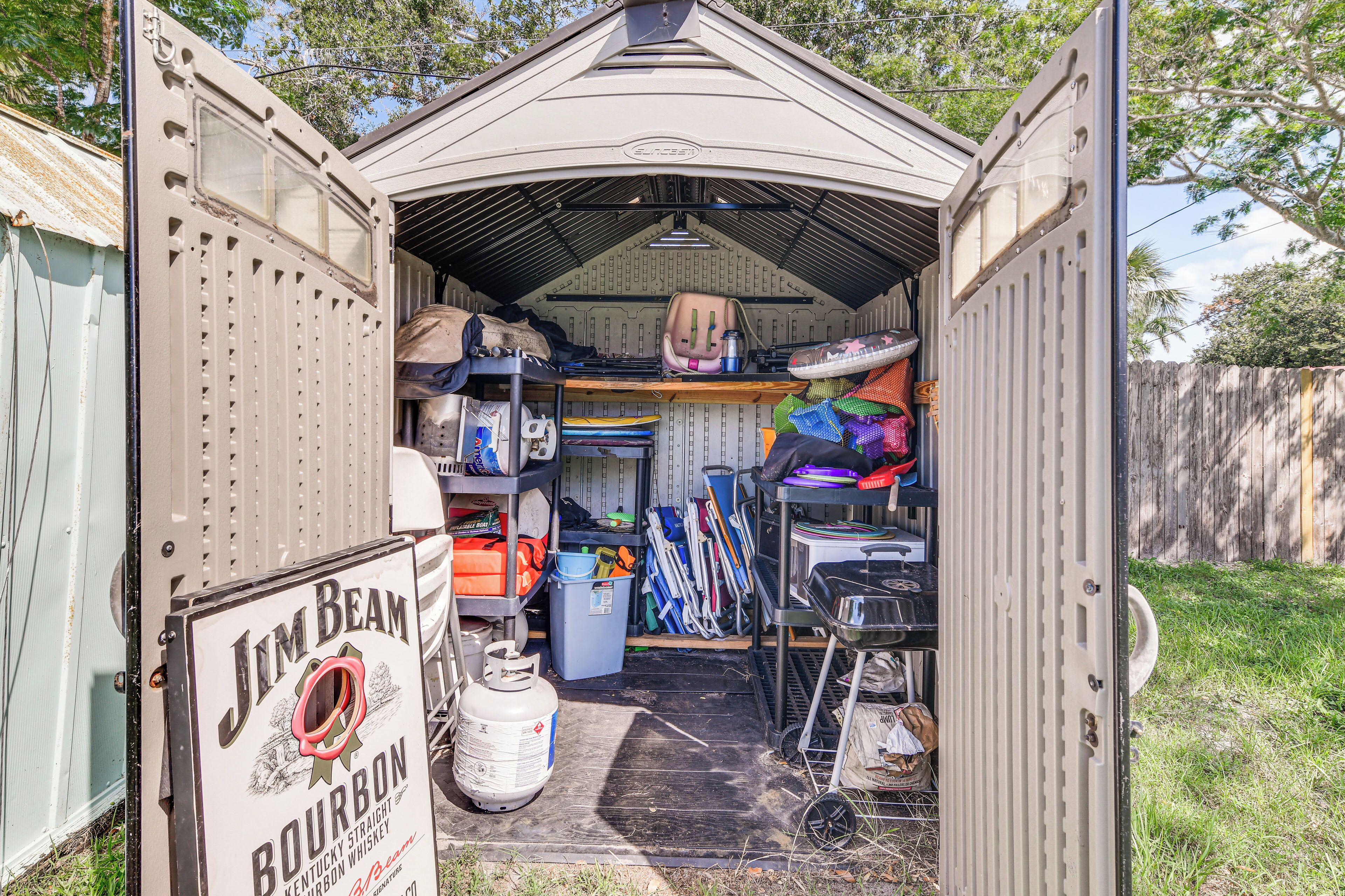 A small, open-sided shed with a metal roof, containing various items like a gas cylinder, tools, and storage bins.