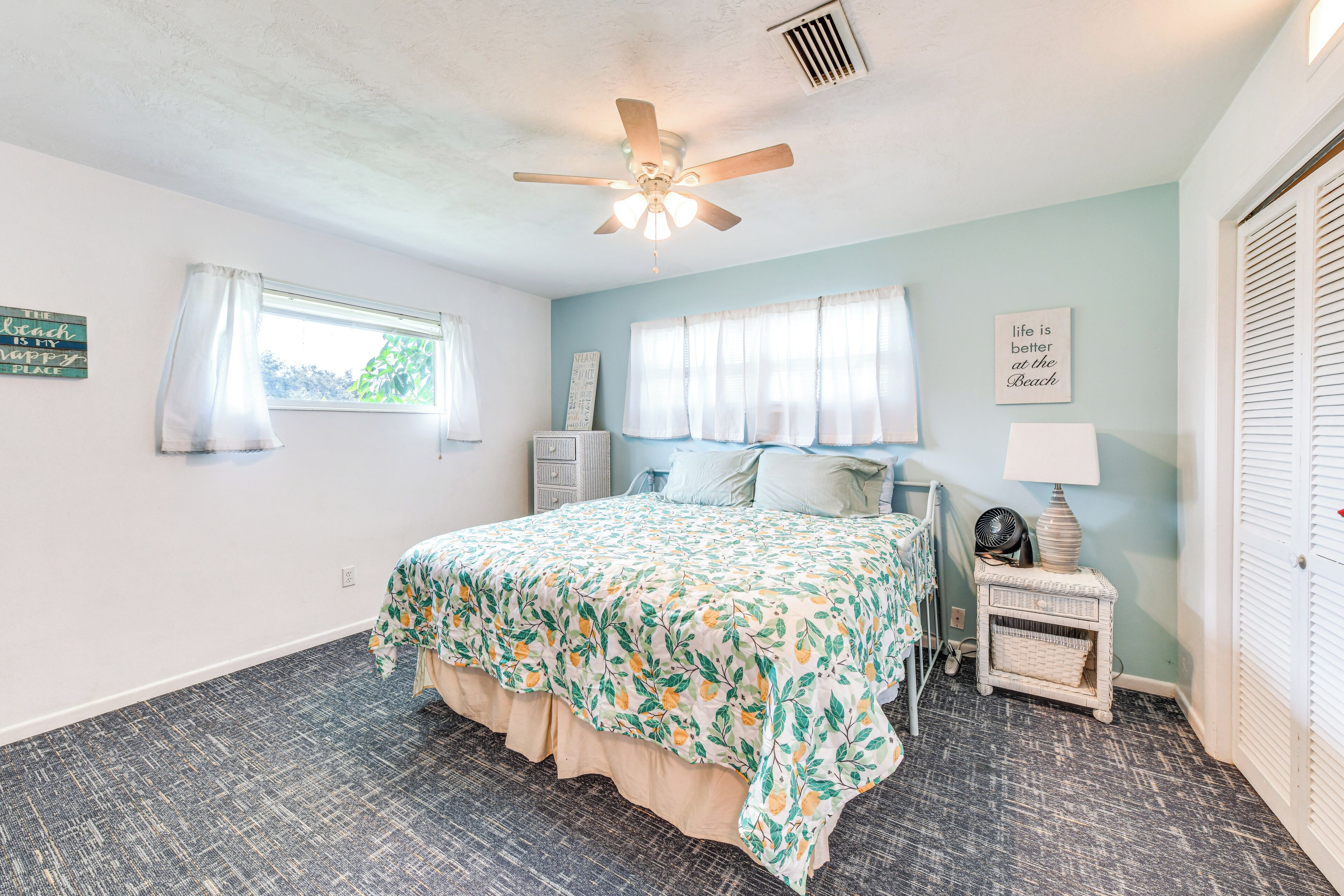 A bedroom with a bed, a ceiling fan, a window with curtains, a bedside table with a lamp, and a wall with a sign.