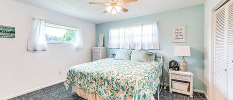 A bedroom with a bed, a ceiling fan, a window with curtains, a bedside table with a lamp, and a wall with a sign.