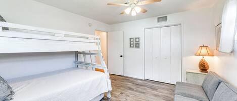 Una habitación con literas, un sofá gris, ventilador de techo, una puerta y una mesita de noche con lámpara.
