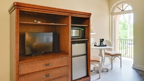 A compact room with a wooden cabinet containing a TV, microwave, and refrigerator. There is a small table with a lamp and a chair. A balcony with a railing and a view of greenery is visible through an arched window. - Poolside Paradise w/ Dock in Everglades! (Naples)