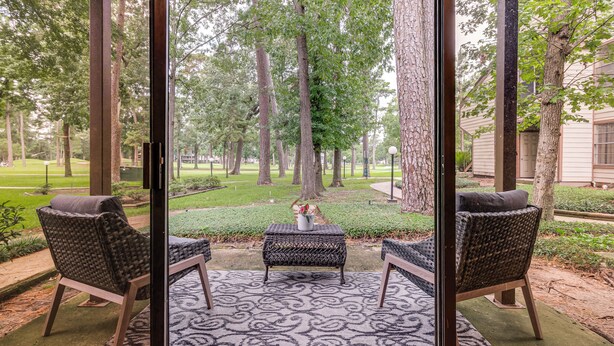 A covered patio with wicker furniture and a patterned rug, overlooking a well-maintained garden and trees. - Walk to Lake Conroe - Condo w/ Community Perks! (Montgomery)