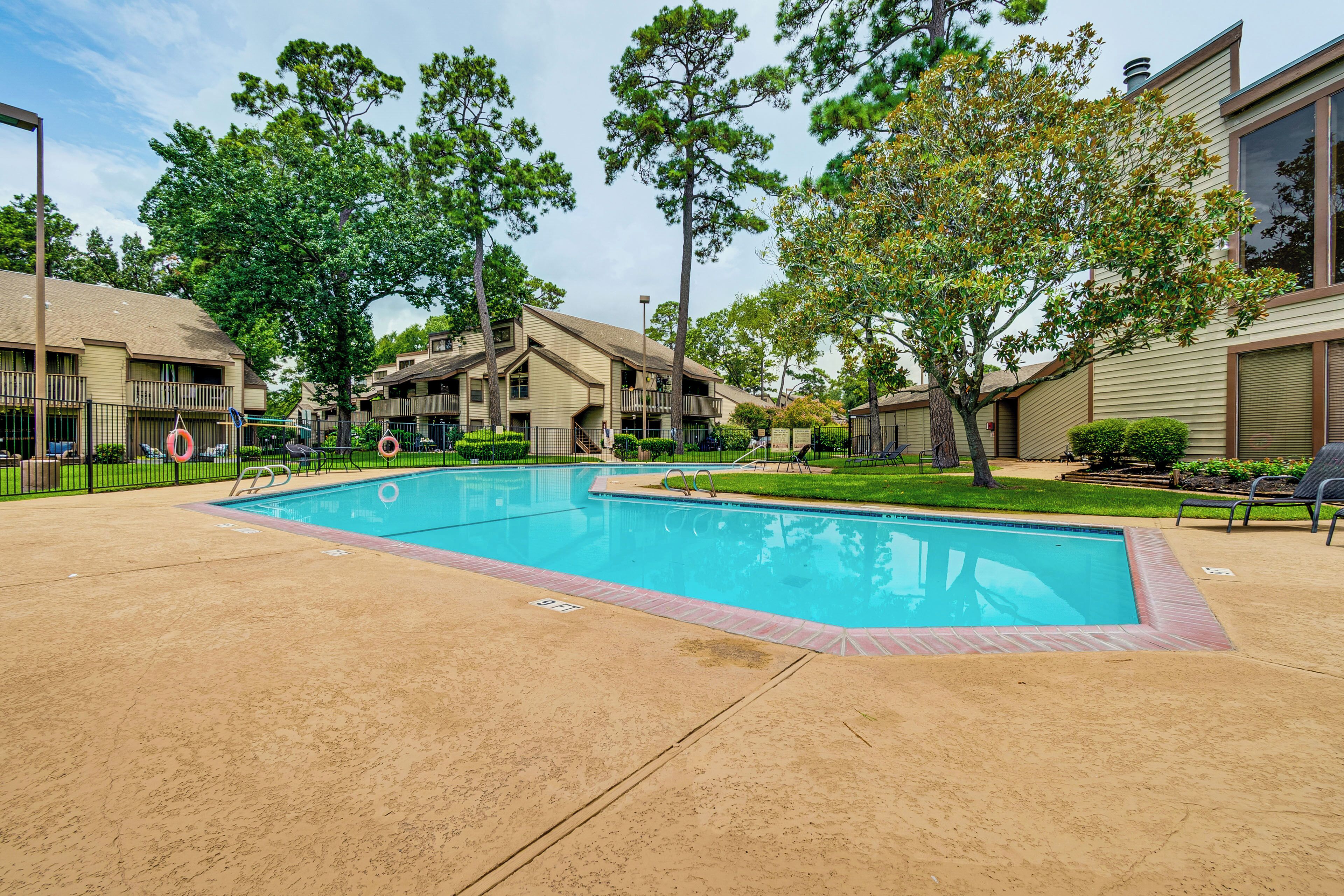 A swimming pool surrounded by a sandy deck, with a building featuring multiple balconies and a large window on the right.