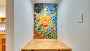 En hörna i ett rum med ett träbord och en stol, en vägg med ett färgstarkt mosaikmönster och en vit vägg med en ljusströmbrytare.