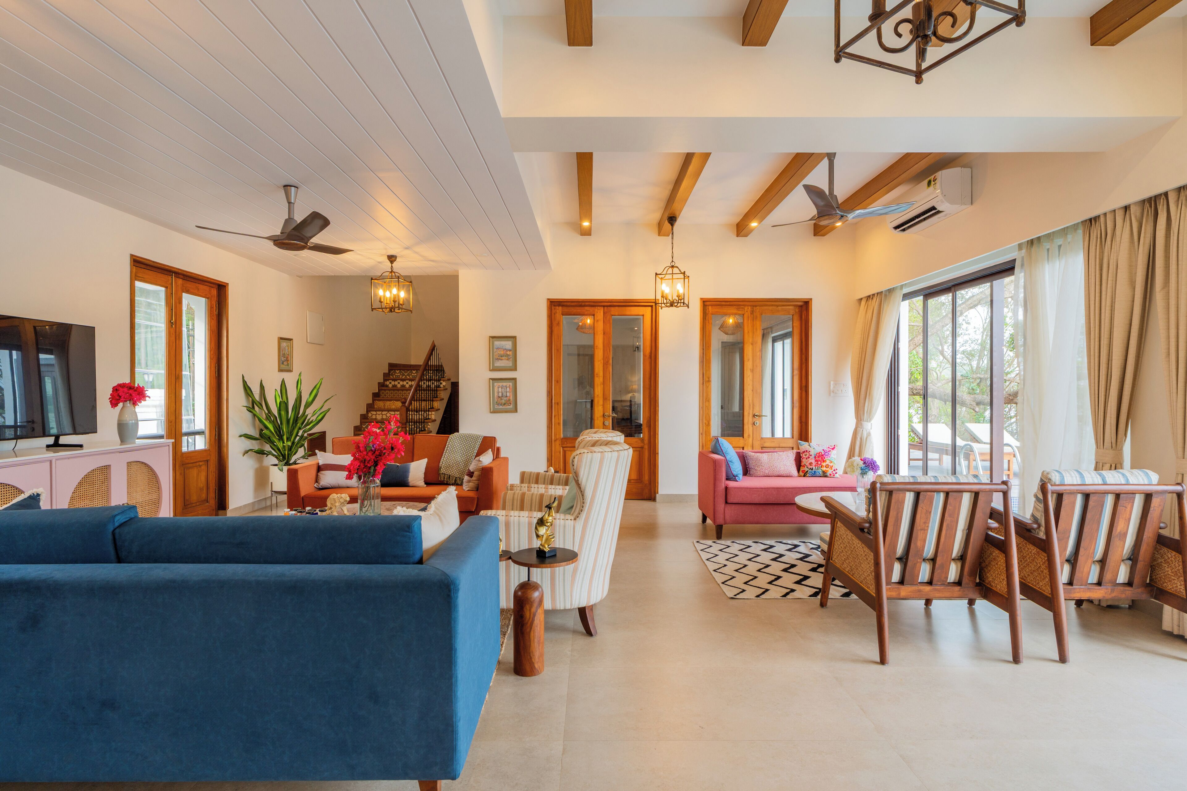 A spacious living area with a blue sofa, striped armchairs, and a pink sofa. There is a staircase leading to another level, a large window with curtains, and a ceiling fan.