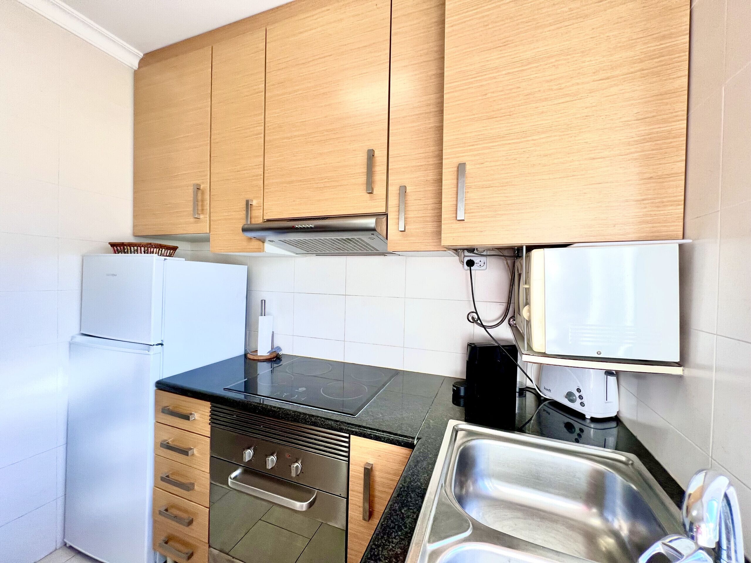 A compact kitchen with wooden cabinets, a built-in oven, a microwave, and a stainless steel sink.